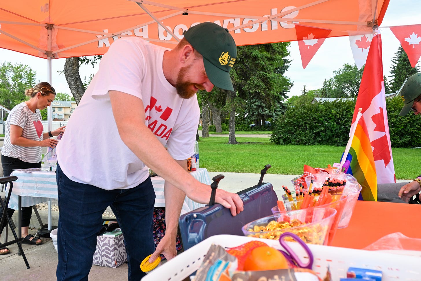 A man in a green hat looks down into a navy blue cooler and holds a small cup of ice cream in his hand under a bright orange canopy tent.
