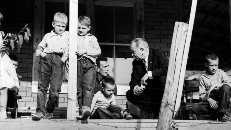 a black and white archival photo shows an older man in a suit and tie stooped down on a front porch to speak with a man and his five children