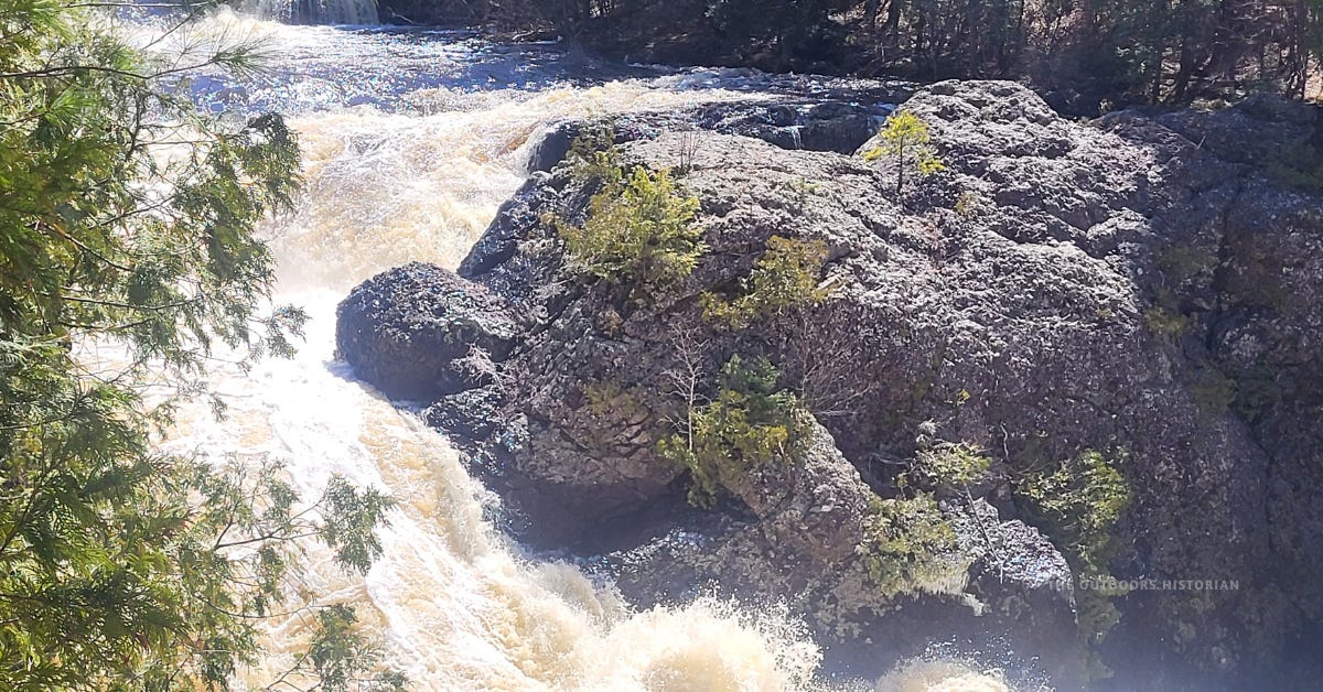 A horizontal view of Upper Potato River Falls at high spring flow. Rushing white water pours over dark, sharply tilted basalt rock layers. Small, scrappy cedar trees cling to the rugged, vertical walls of the gorge.
