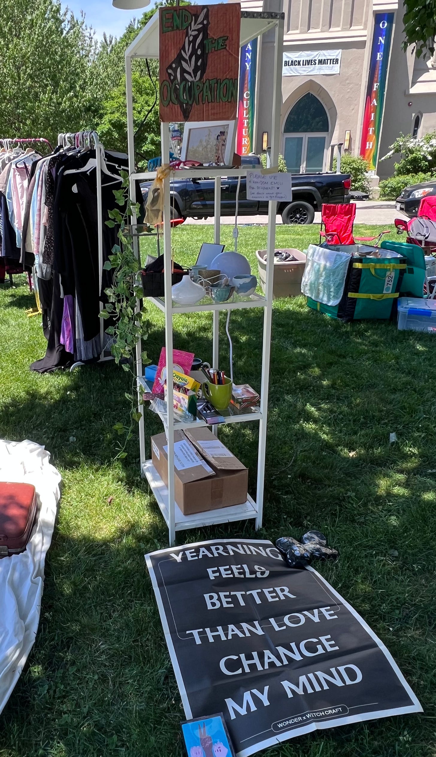 A picture from a recent yard sale for Palestine with a few clothing racks on a church lawn, a bookshelf, and a paper poster weighted down on the grass. Poster is black with white letters and reads “Yearning is better than loving change my mind” with “Wonder x Witchcraft” on the bottom of the poster.