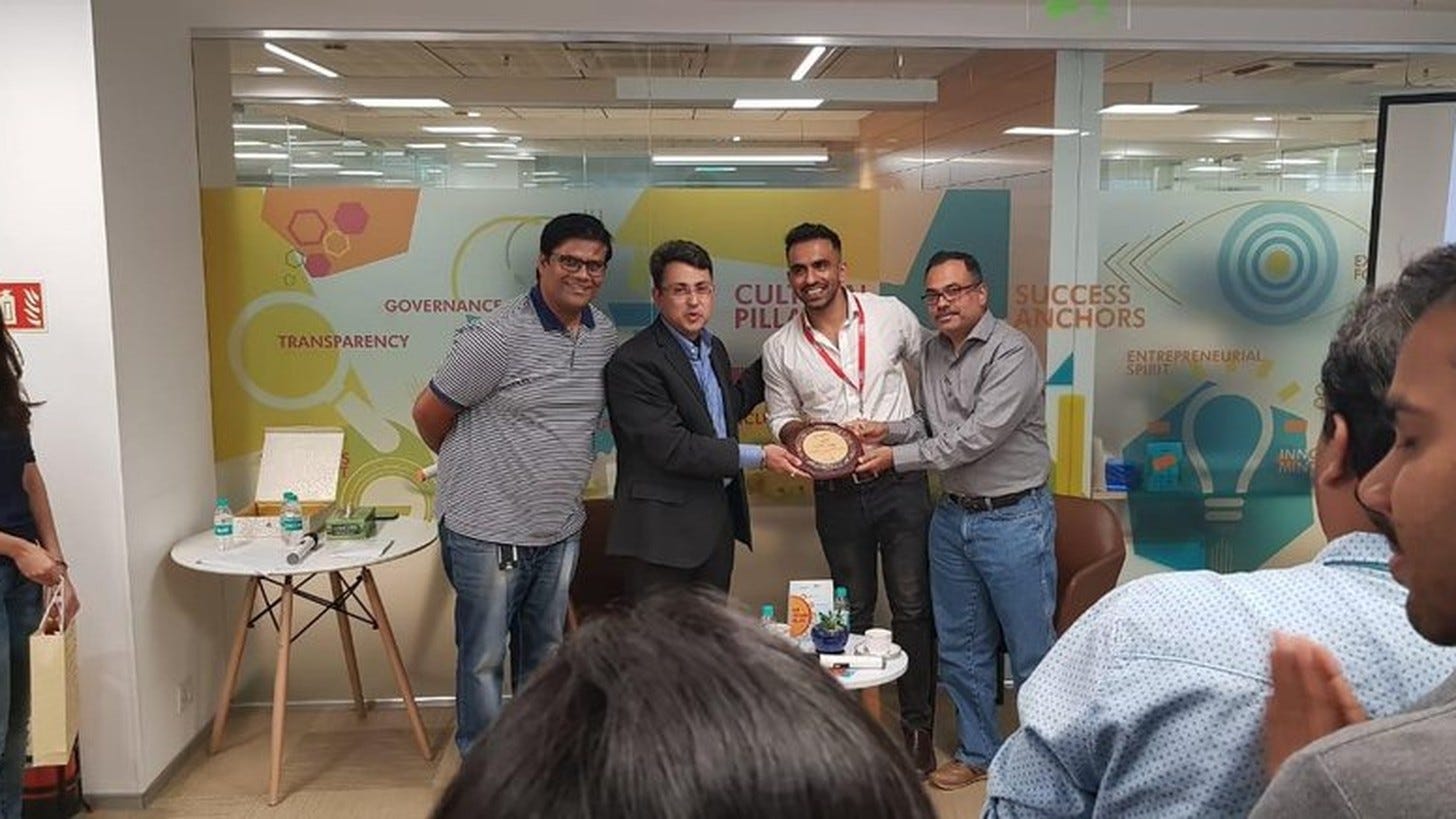 Avi holding a plaque with three colleagues at an office award ceremony, people clapping in the foreground.