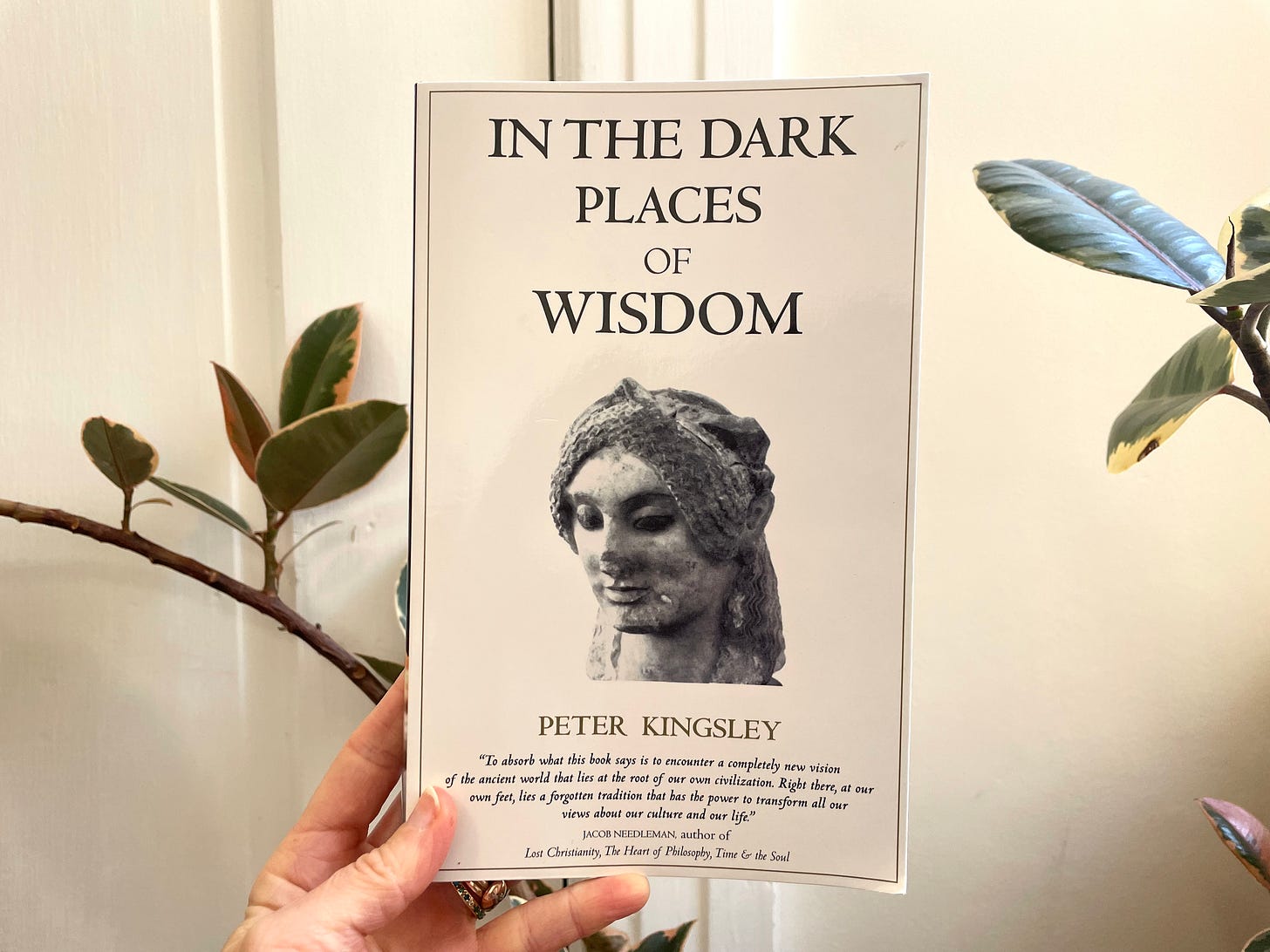 image of Casey holding up "In the Dark Places of Wisdom" by Peter Kingsley. White cover with an ancient bust photo on the cover.