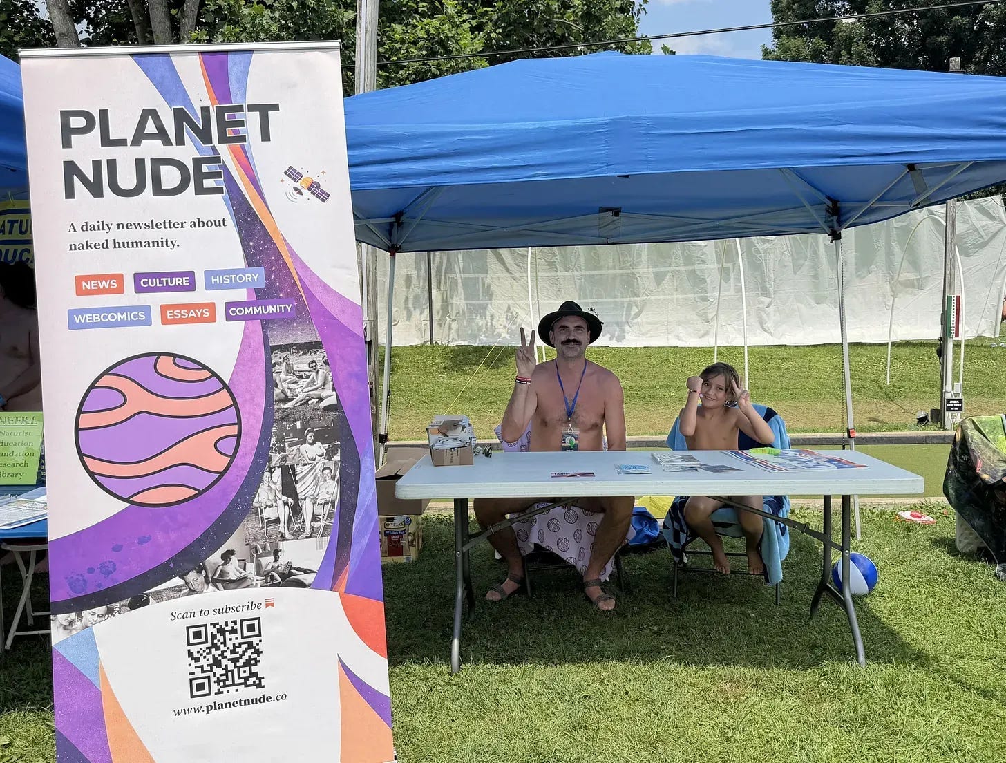 Evan Nix and his young son sit behind a folding table at an outdoor vendor booth, next to a tall Planet Nude banner, both making peace signs under a blue canopy tent. Evan Nix and his young son sit behind a folding table at an outdoor vendor booth, next to a tall Planet Nude banner, both making peace signs under a blue canopy tent.