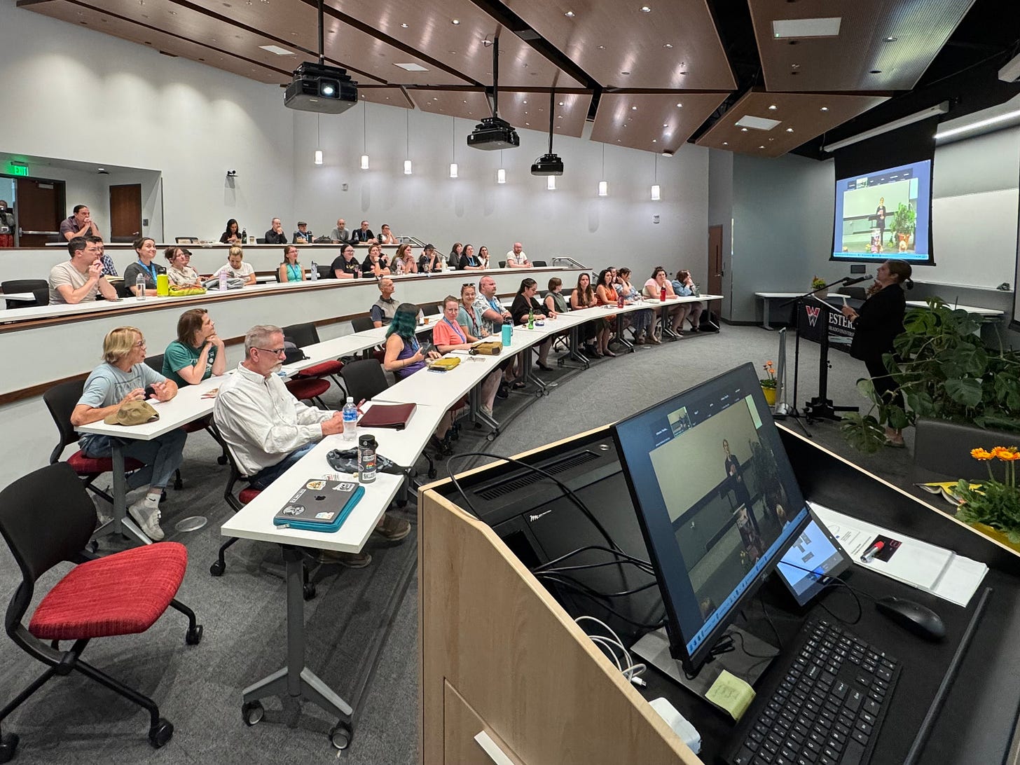 High-ceilinged auditorium with four rows of seats mostly filled with people, with a person at a podium in front with a TV monitor