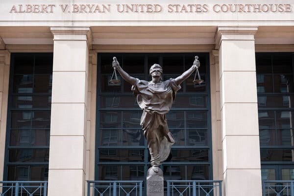 A statue of blindfolded Justice with outstretched arms holding a pair of scales in front of the squared columns of a building. 