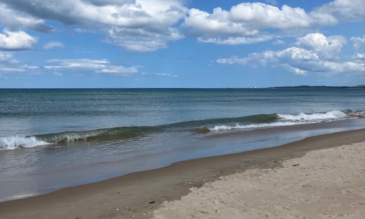 A tranquil view of the north Aberdeenshire coast, showing gentle waves rolling onto a sandy beach under a bright blue sky scattered with fluffy white clouds. The distant shoreline is faintly visible on the horizon.