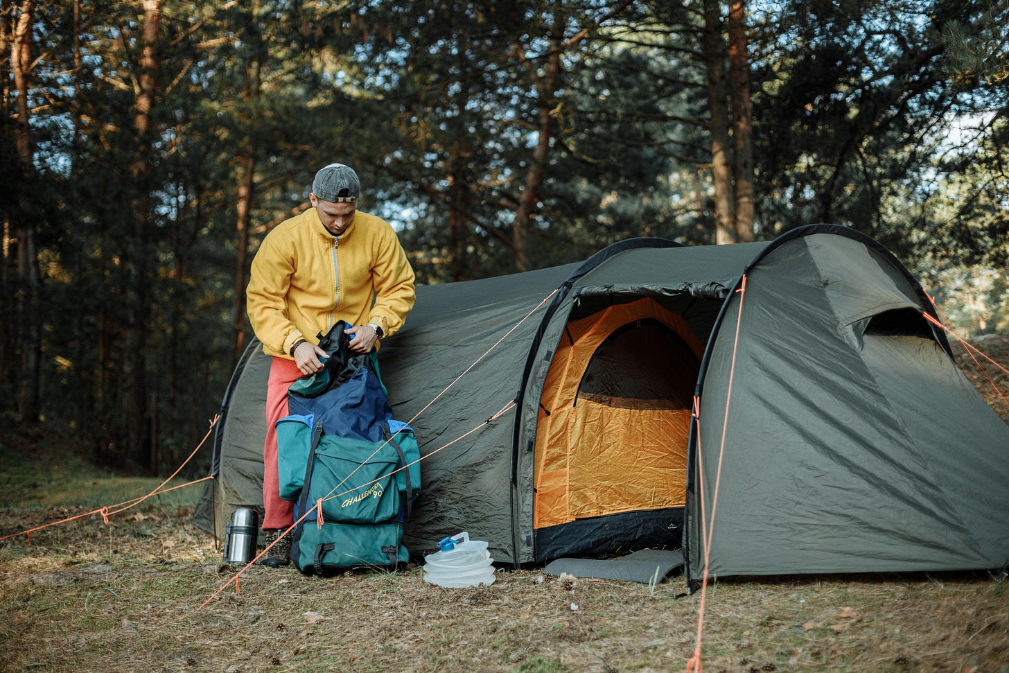 A person in a yellow jacket and cap is setting up or packing gear outside a large green camping tent in a forested area. The tent has an orange interior, and various camping items, including a backpack, thermos, and water container, are spread out on the ground nearby. Sunlight filters through the trees, creating a peaceful outdoor setting. A person in a yellow jacket and cap is setting up or packing gear outside a large green camping tent in a forested area. The tent has an orange interior, and various camping items, including a backpack, thermos, and water container, are spread out on the ground nearby. Sunlight filters through the trees, creating a peaceful outdoor setting.