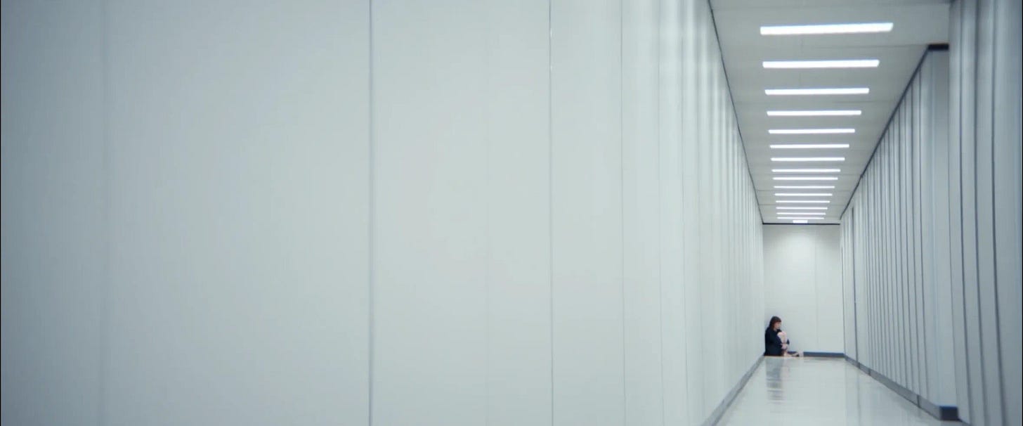 A woman, Hally, from the TV show Severence, sits in a distant corner of an all-white corridor.