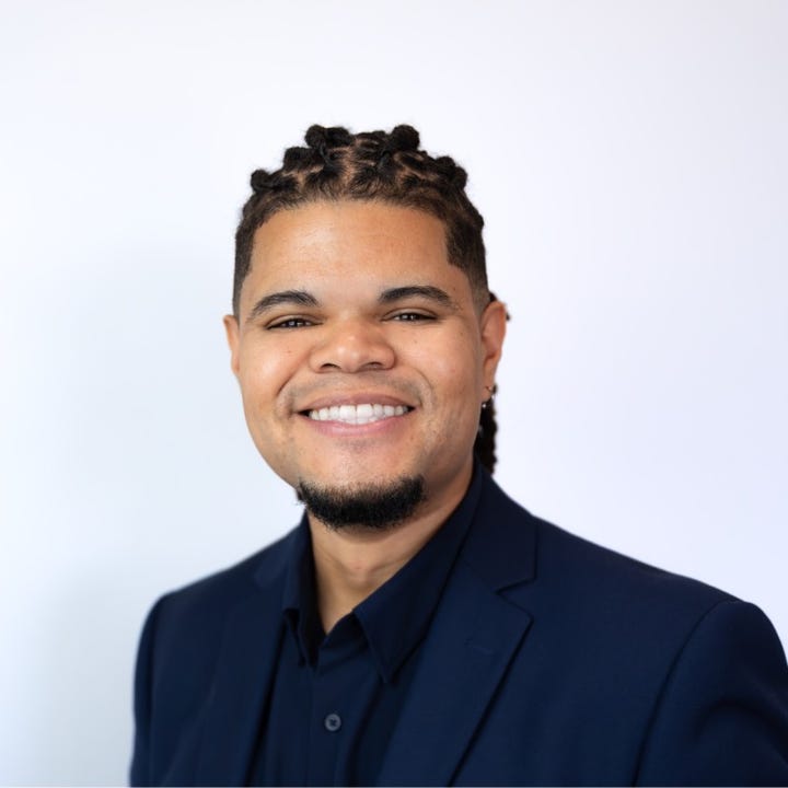 Chris Jones, a man with a friendly smile and braided hair, standing against a light background wearing a blue suit jacket and a black polo