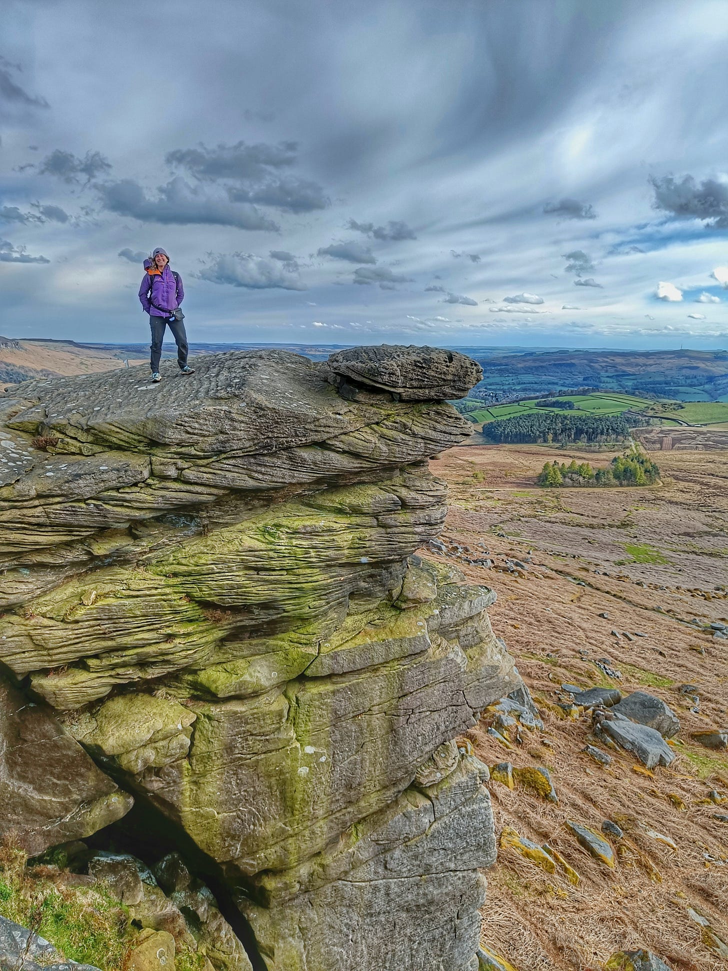 A woman stands on top of Stanage Edge, with a view of the Peak District National Park in the background