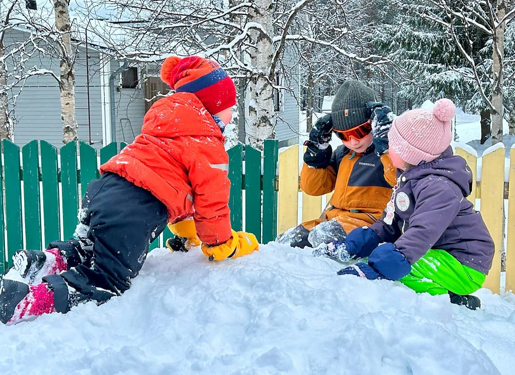 Three children in winter clothing play on a mound of snow in a snowy yard, with a green-and-yellow fence and snow-covered trees behind them.