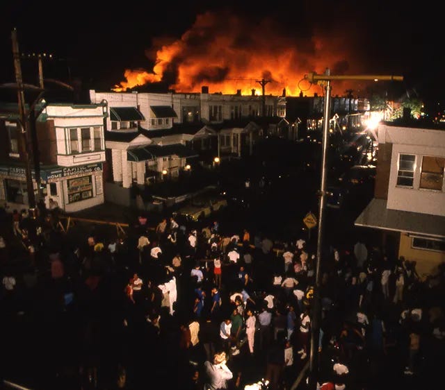 A stunned crowd watches in horror as an entire row of buildings goes up in flames in Philadelphia, following the tragic May 13, 1985 bombing. A stunned crowd watches in horror as an entire row of buildings goes up in flames in Philadelphia, following the tragic May 13, 1985 bombing.