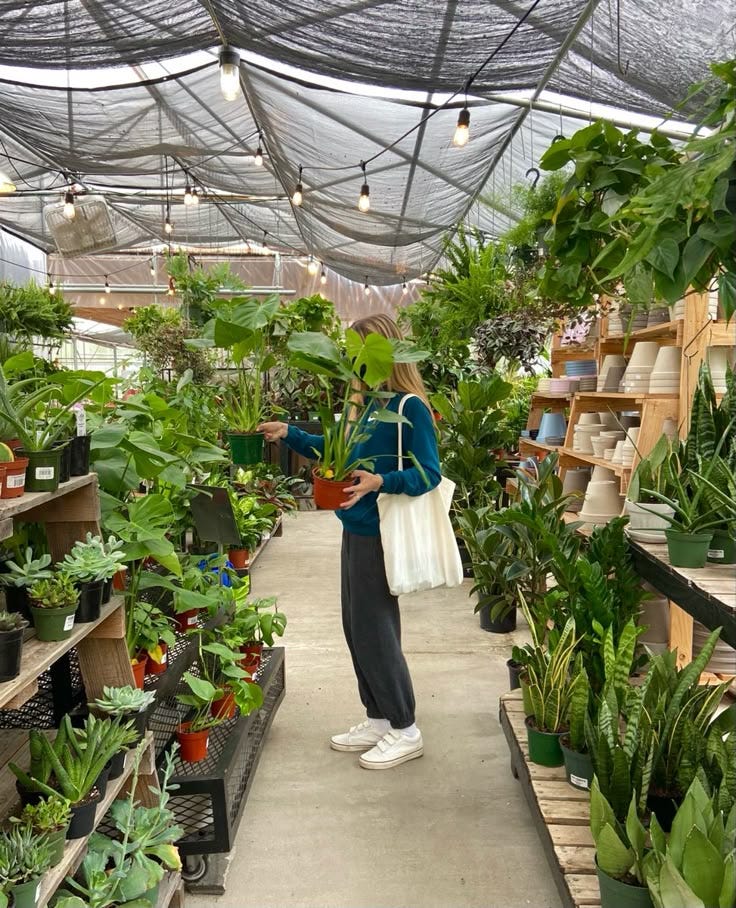 This may contain: a woman is looking at plants in a greenhouse with lots of potted plants on the shelves