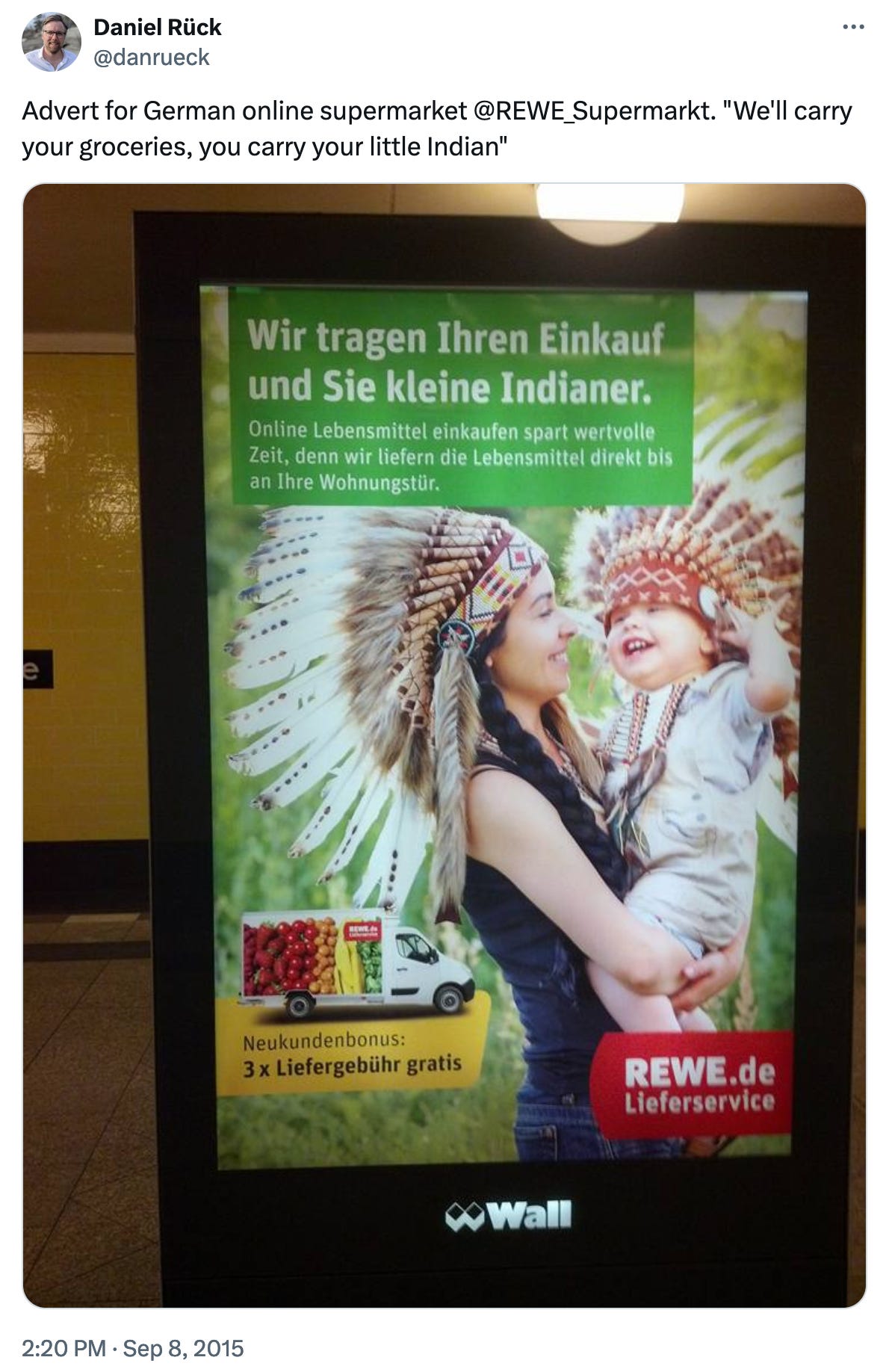 A picture of an ad in a German subway, showing a woman and a child, both wearing headdresses. The ad is for a German grocery company's delivery service, and in German reads: "We'll take your groceries, you take your little Indian."