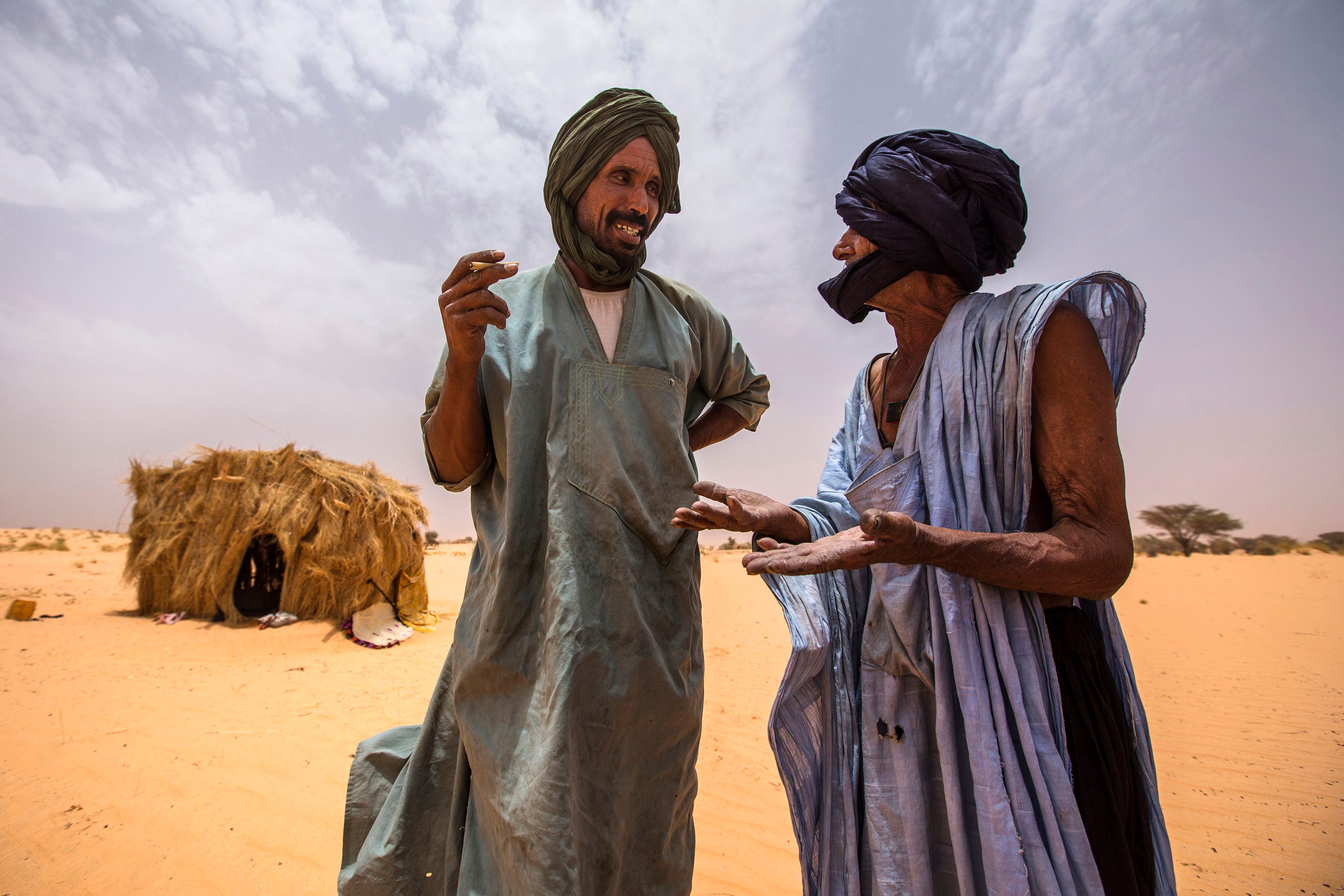 Two men in traditional robes and head coverings talk and gesture in a sandy desert landscape, with a straw hut and cloudy sky in the background.