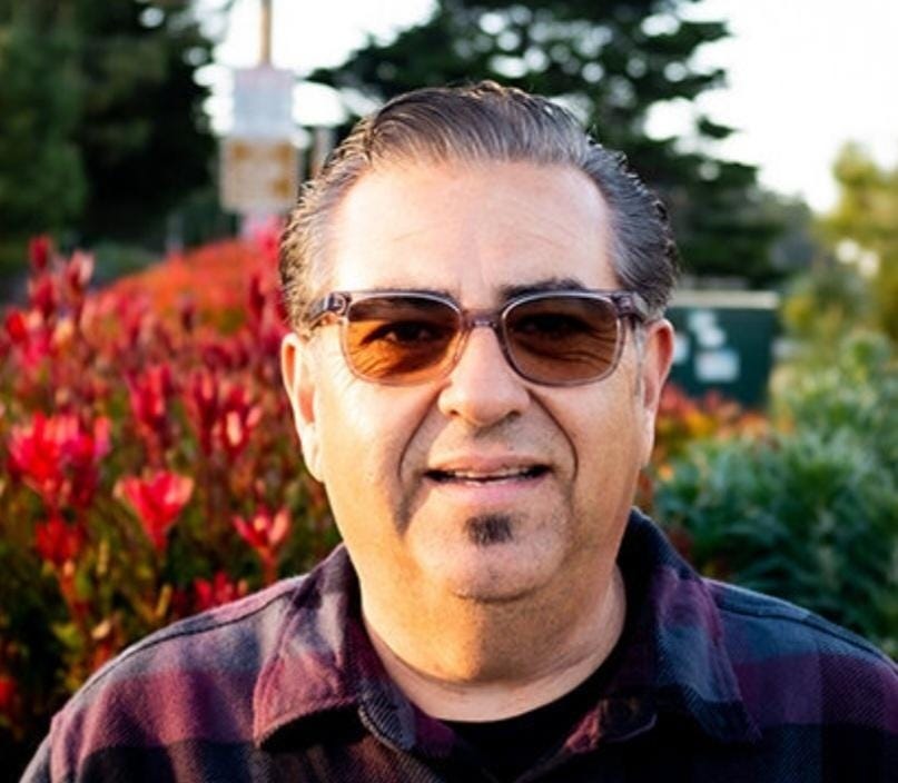 A middle aged man in tinted glasses is smiling at the camera. He's outside, with big red flowers behind him.