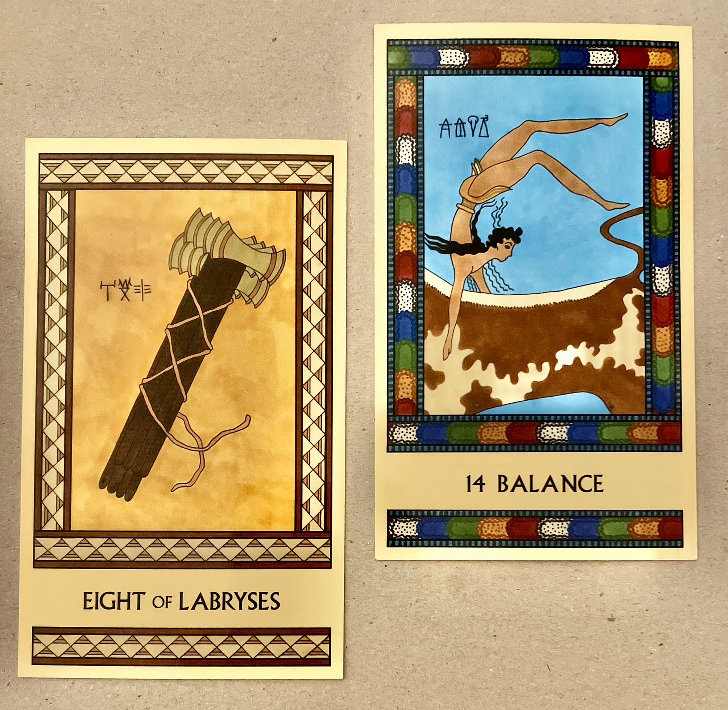 Two Minoan Tarot cards side by side on a dark beige surface. The Eight of Labryses is painted in shades of deep yellow, cream, and brown. It shows a group of eight long-handled Labryses apparently bound together with rope, but the ends of the rope are loose, so the labryses can be removed. The Balance card has a bright multicolor border. It shows a Minoan bull leaper balancing upside down on the back of a brown-and-cream spotted bull, with a light blue background behind the scene. 