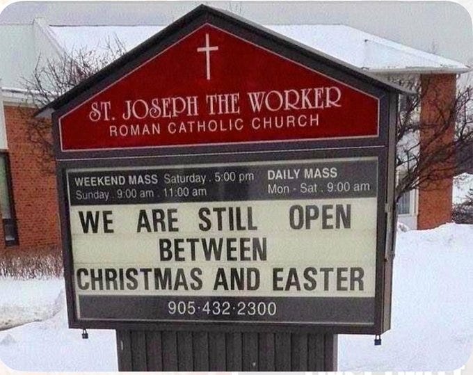 A sign for St. Joseph the Worker Roman Catholic Church. The sign has a red triangular top with a cross and text reading "St. Joseph the Worker Roman Catholic Church." Below, a black board displays text: "WE ARE STILL OPEN BETWEEN CHRISTMAS AND EASTER" and mass times, including "WEEKEND MASS Saturday 5:00 pm, Sunday 9:00 am, 11:00 am" and "DAILY MASS Mon. - Sat. 9:00 am, Open." The background shows a snowy landscape and a brick building.