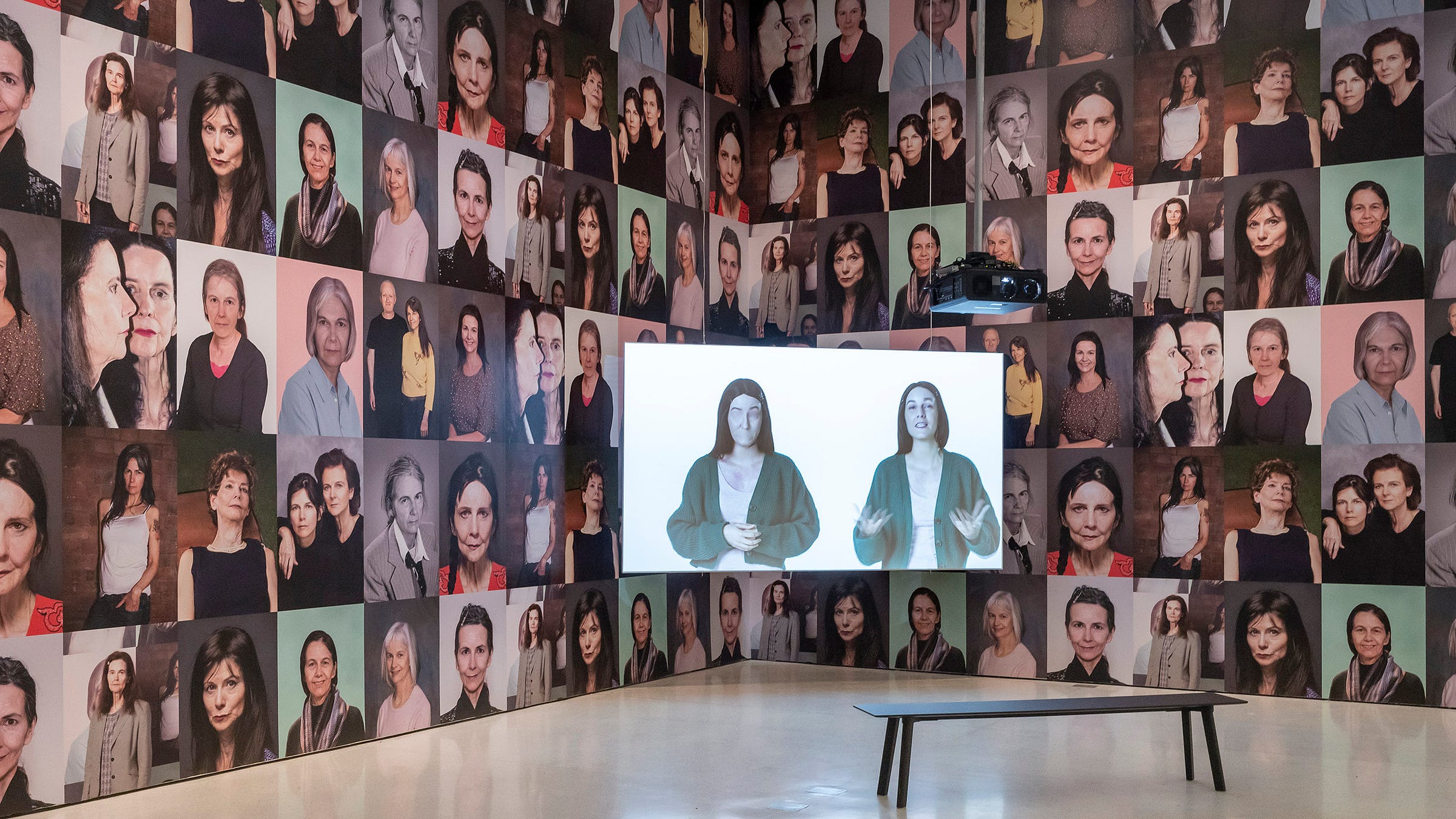Uncanny portraits of different women cover two walls. In front of the corner where the walls meet is a screen with a projection of a woman standing next to a version of herself, but the version is missing facial features.