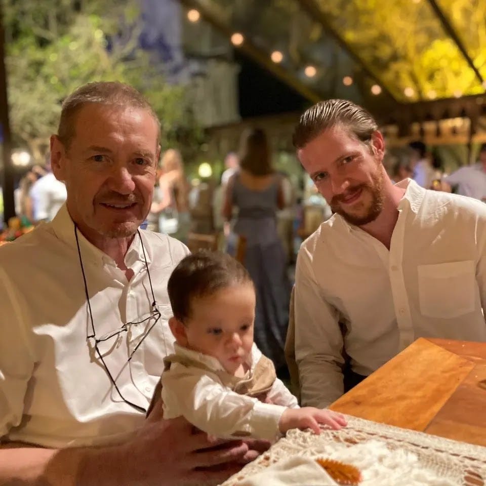 Two adult white men dressed formally smile at the camera, one of them holds a baby