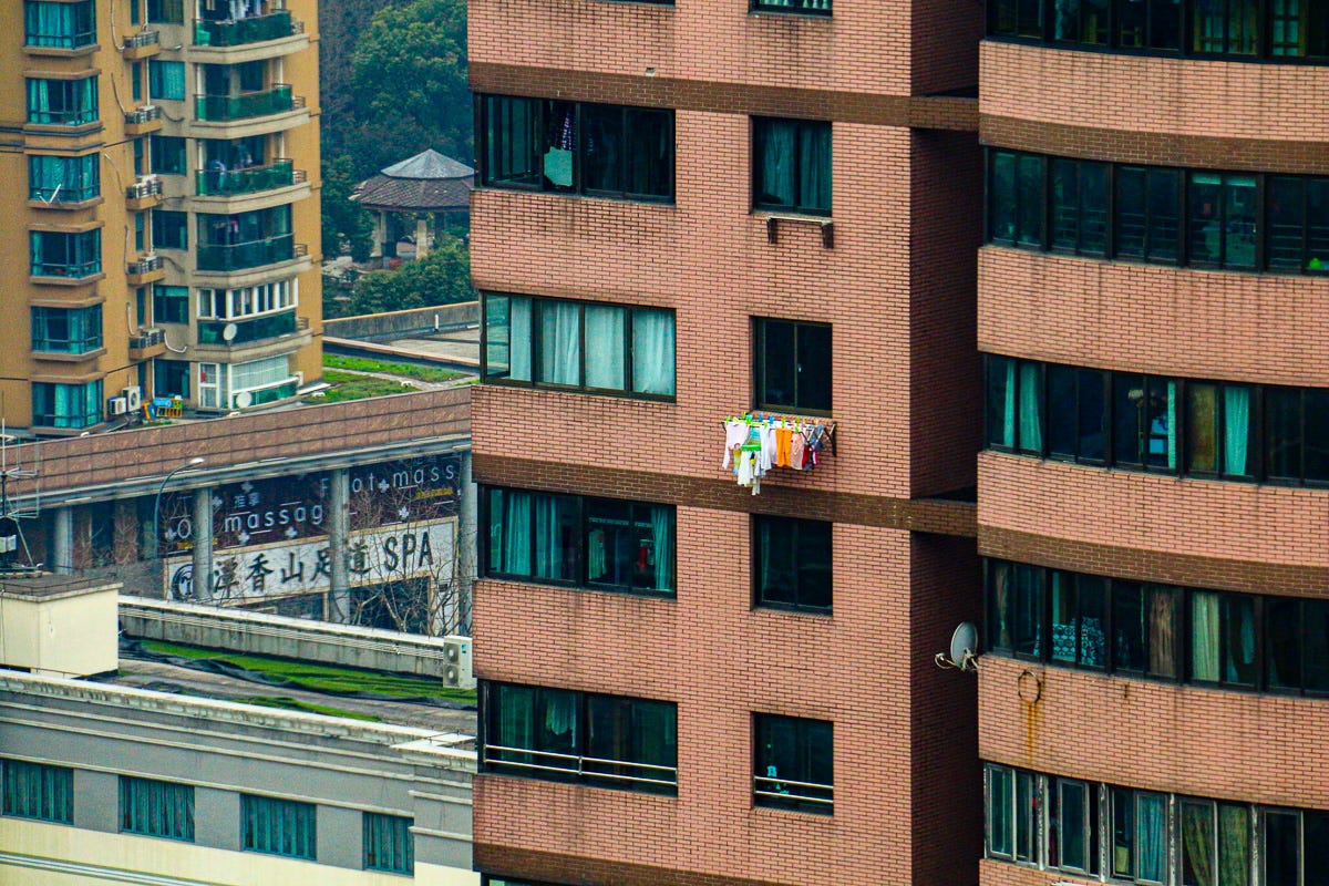A compact balcony protrudes from a high-rise with children's clothes drying on a rack. Laundry spills out into the urban skyline of Shanghaiâ€™s residential towers.