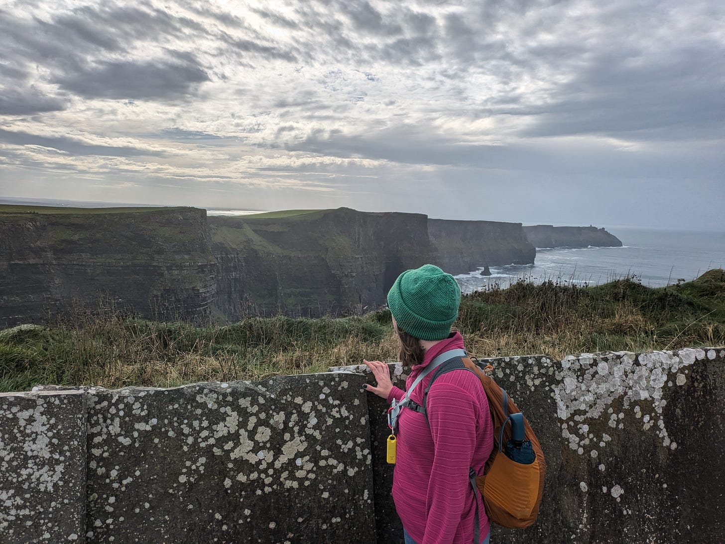 White woman stand with her back to the camera wearing a green hat, pink fleece, and orange backpack looking out at the Cliffs of Moher on a partially cloudy day