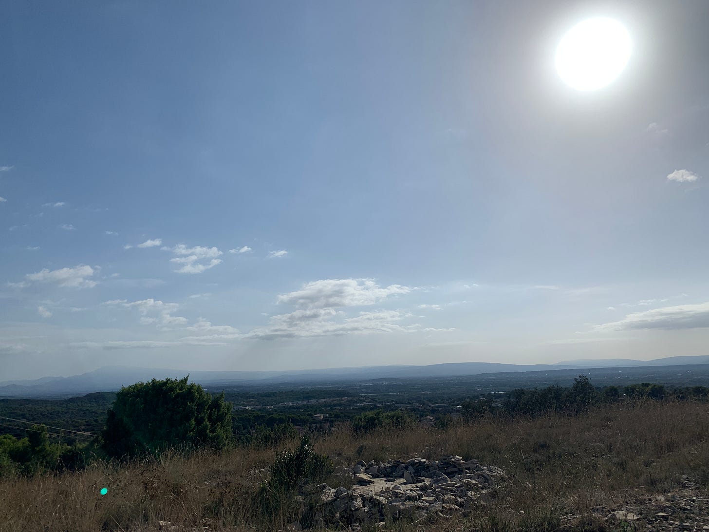 Vue panoramique d'une colline baignée par le soleil en plein après-midi, avec un ciel bleu clair et quelques nuages flottant à l'horizon. Des arbres verts épars et un amas de pierres sont visibles au premier plan, tandis que des collines boisées s'étendent au loin.