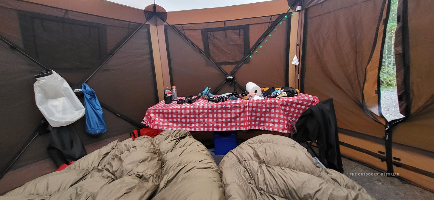 A view from the head of a sleeping cot looking down into a cozy tent interior. Two heavy, dark green 0-degree sleeping bags are laid out, and small green battery-powered lights are clipped to the tent wall for a soft interior glow.