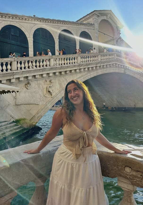 A woman smiles while leaning on a stone railing beside a canal in Venice, with the crowded Rialto Bridge in the background.