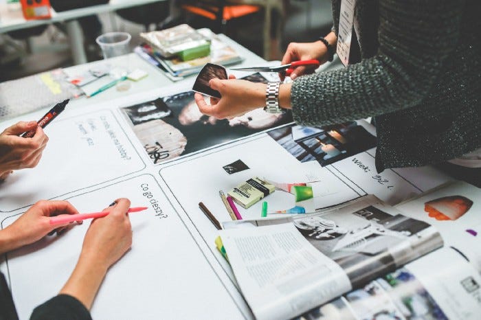 A group of people gathered around the table. Some have markers that are trying to fill in blank white spaces, while another is holding a picture and a pair of scissors to try and place images down on the board. A group of people gathered around the table. Some have markers that are trying to fill in blank white spaces, while another is holding a picture and a pair of scissors to try and place images down on the board.