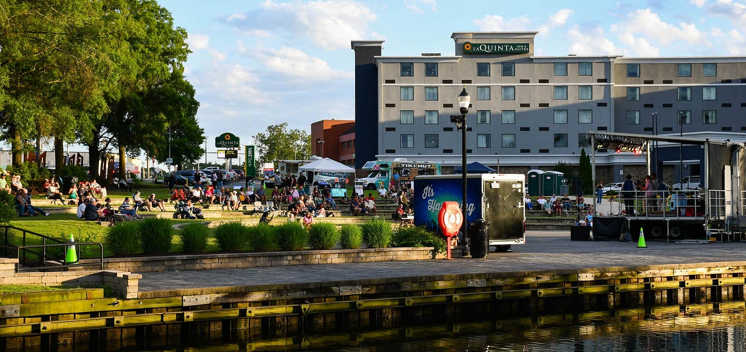 A lively cityscape featuring pedestrians walking by the water, with urban architecture in the background.