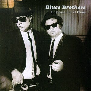 Album cover of Briefcase Full Of Blues that features two men dressed in classic blues attire: dark suits, white shirts, skinny black ties, and black hats. Both wear dark sunglasses and have a serious, cool expression. Text in the upper right corner reads: "Blues Brothers Briefcase Full of Blues." Picture Credit: Wikipedia