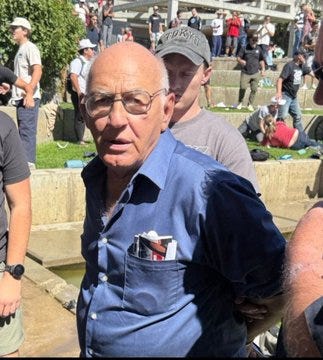 Ένας άντρας που φοράει γυαλιά και ένα μπλε πουκάμισο με κουμπιά με τσέπη, στέκεται έξω ανάμεσα σε ένα πλήθος. Άνθρωποι με casual ρούχα είναι ορατοί στο βάθος, μερικοί κάθονται σε τσιμεντένια σκαλοπάτια και γρασίδι. Το σκηνικό φαίνεται να είναι μια δημόσια εκδήλωση ή συγκέντρωση με δέντρα και μια ηλιόλουστη μέρα.