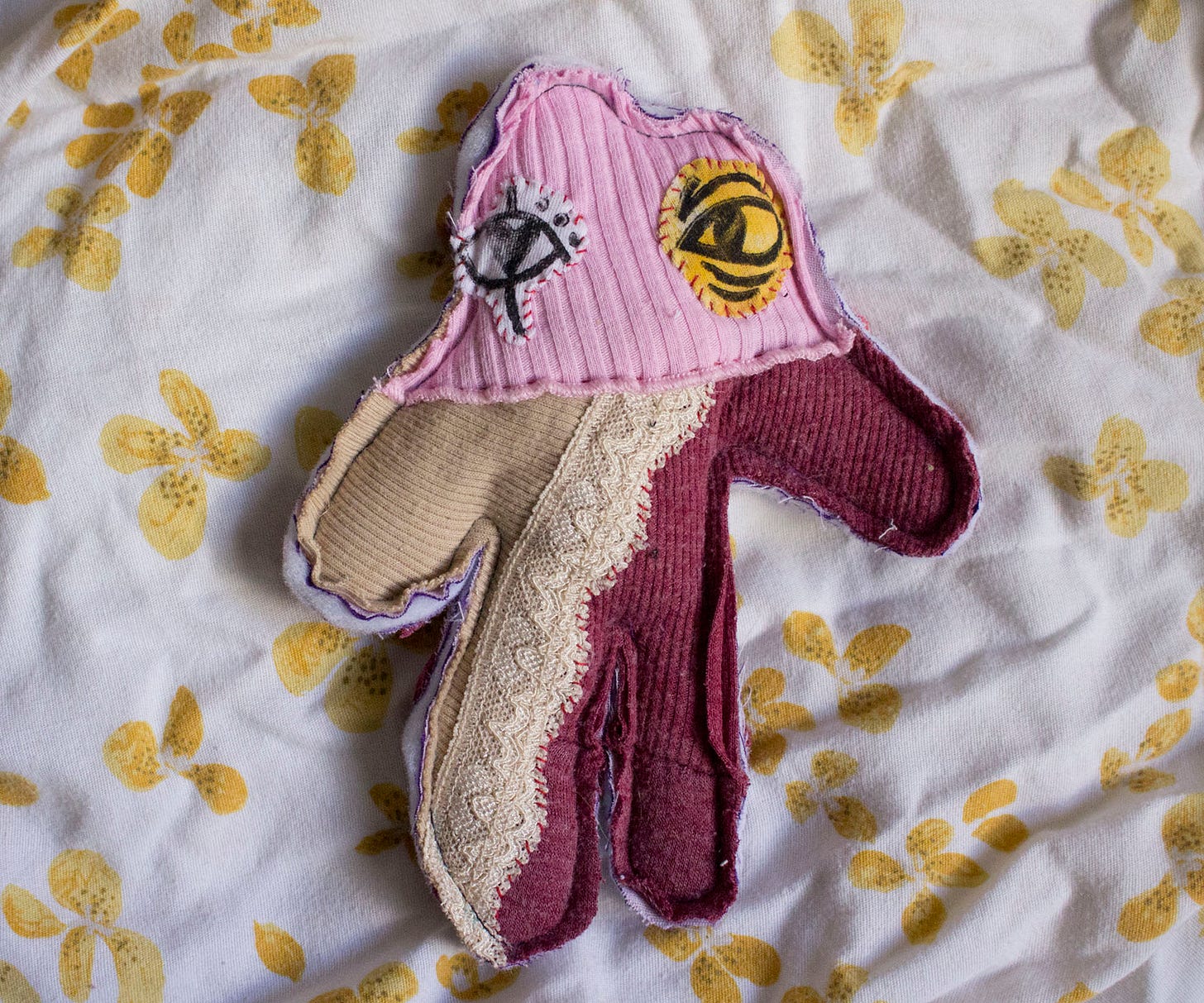 A humanoid creature sewn from red, beige, and pink fabrics. It has mismatched eyes and lace details.