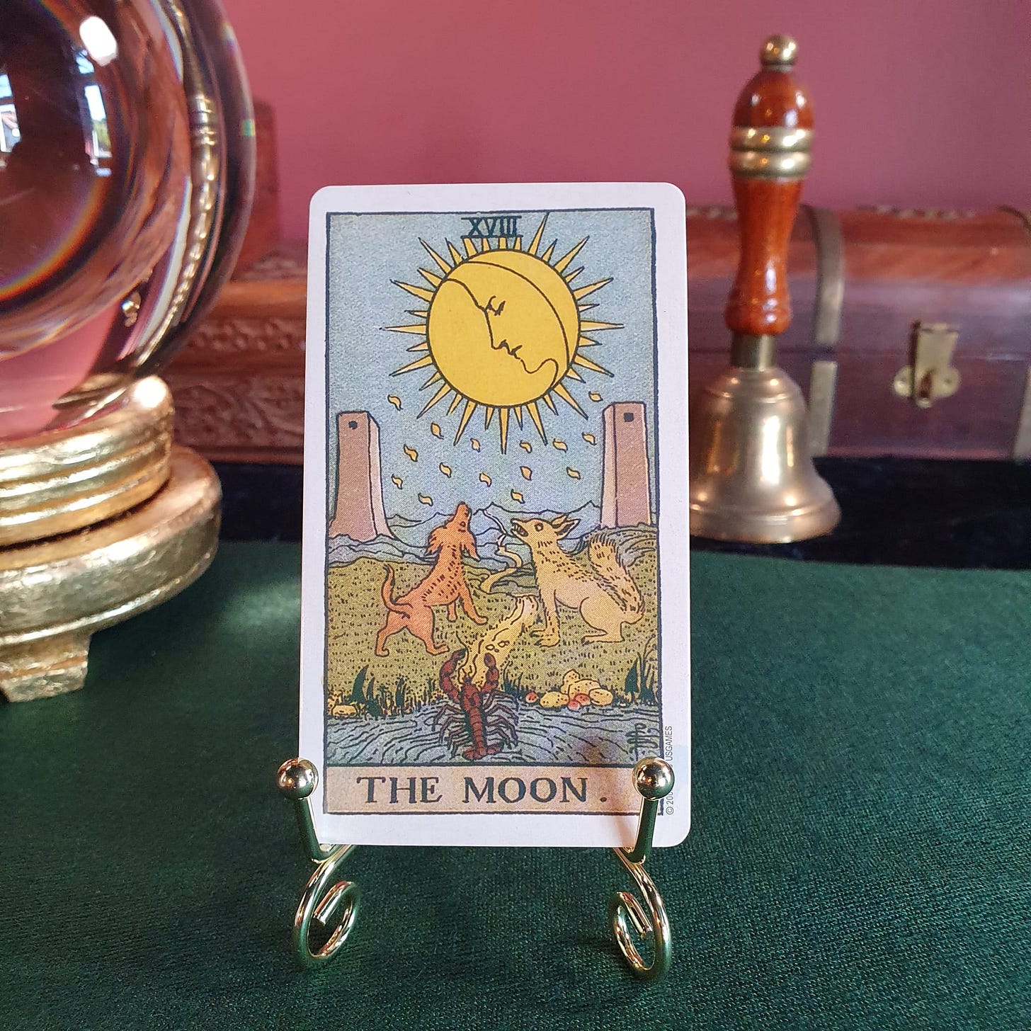 The Moon tarot card from the Smith-Waite deck displayed on a small gold stand, showing a moonlit scene with two towers, two animals, and a crayfish emerging from water.