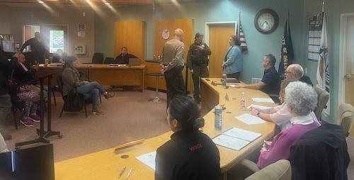 Two uniformed officers are talking to a woman standing at the end of a curved table with several other people in the room watching what is happening.