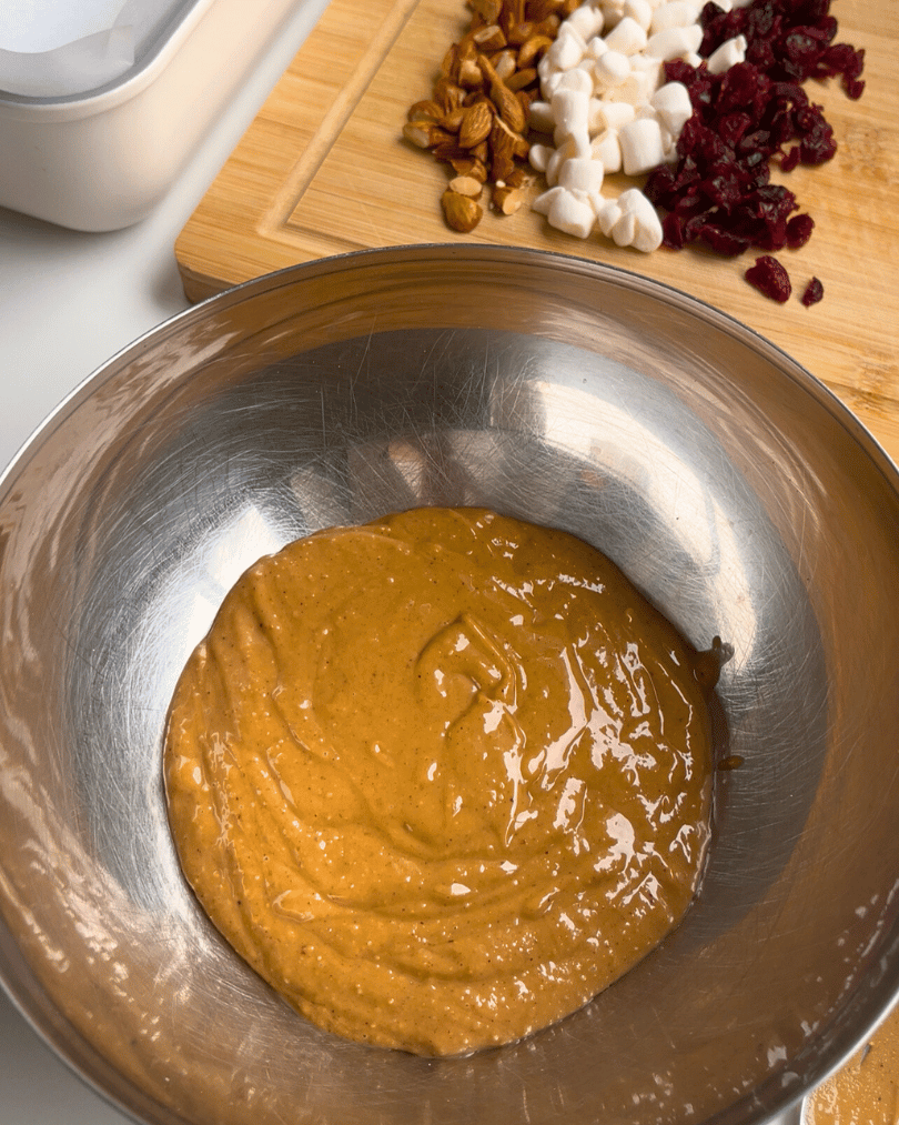 peanut butter in a metal bowl with chopped almond, marshmallows, and dried cranberries on a wooden cutting board behind it