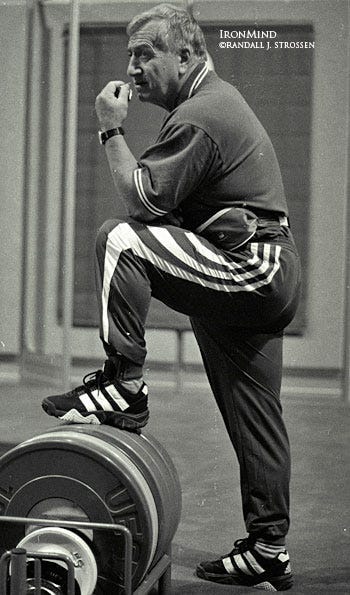Ivan Abadjiev, adorned in Adidas striped track suit and shoes, stands with one foot on a rack of bumper plates for weightlifting, contemplative Ivan Abadjiev, adorned in Adidas striped track suit and shoes, stands with one foot on a rack of bumper plates for weightlifting, contemplative