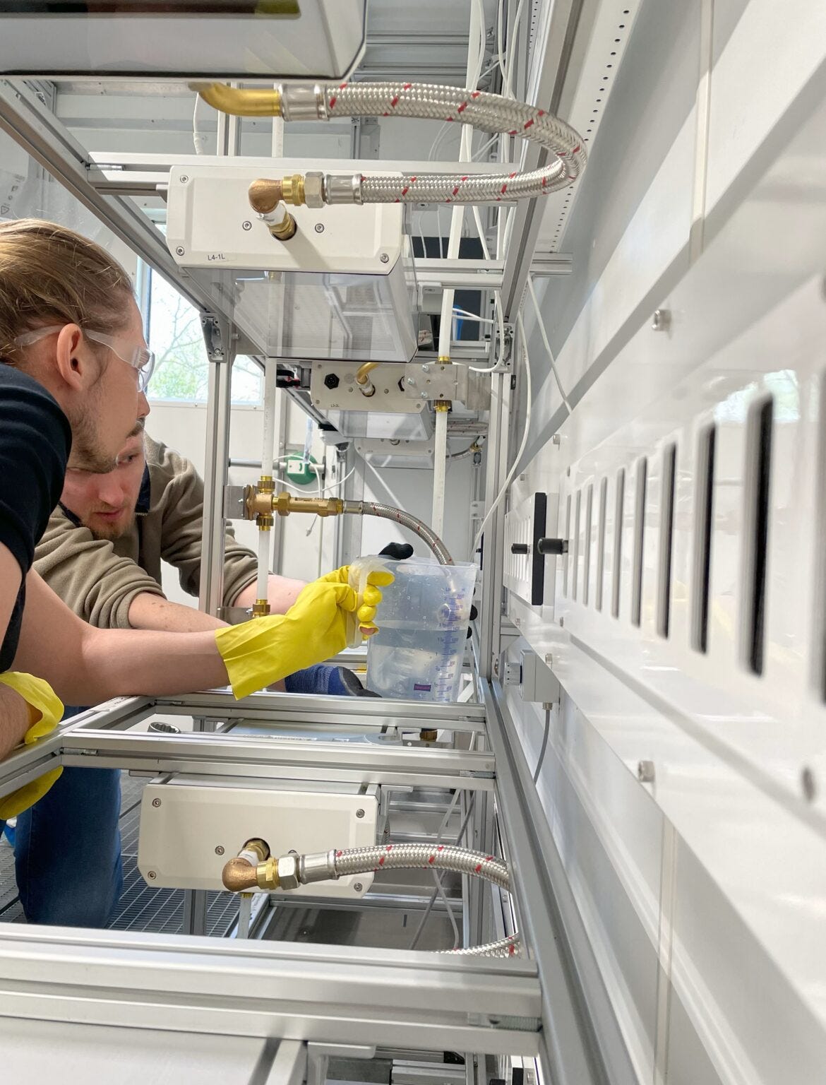 Two men are reaching into a rack of equipment. One is wearing bright yellow rubber gloves and holding a jug half-filled with water under a pipe. They are conducting a leakage test. Two men are reaching into a rack of equipment. One is wearing bright yellow rubber gloves and holding a jug half-filled with water under a pipe. They are conducting a leakage test.