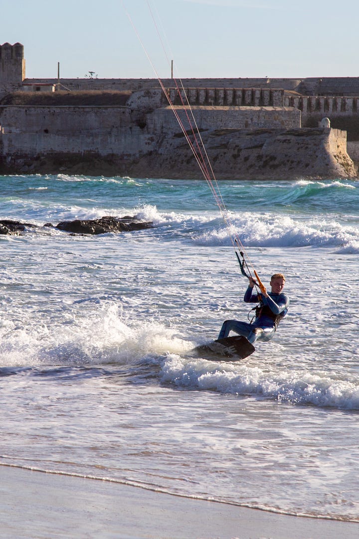 Kitesurfer riding near the medieval castle walls in Tarifa; the Mar Mediterráneo and Océano Atlántico signs marking where two seas meet at the Tarifa causeway; a kitesurfer suspended mid-air above the Strait of Gibraltar; a man running into the surf with a kiteboard on Tarifa beach