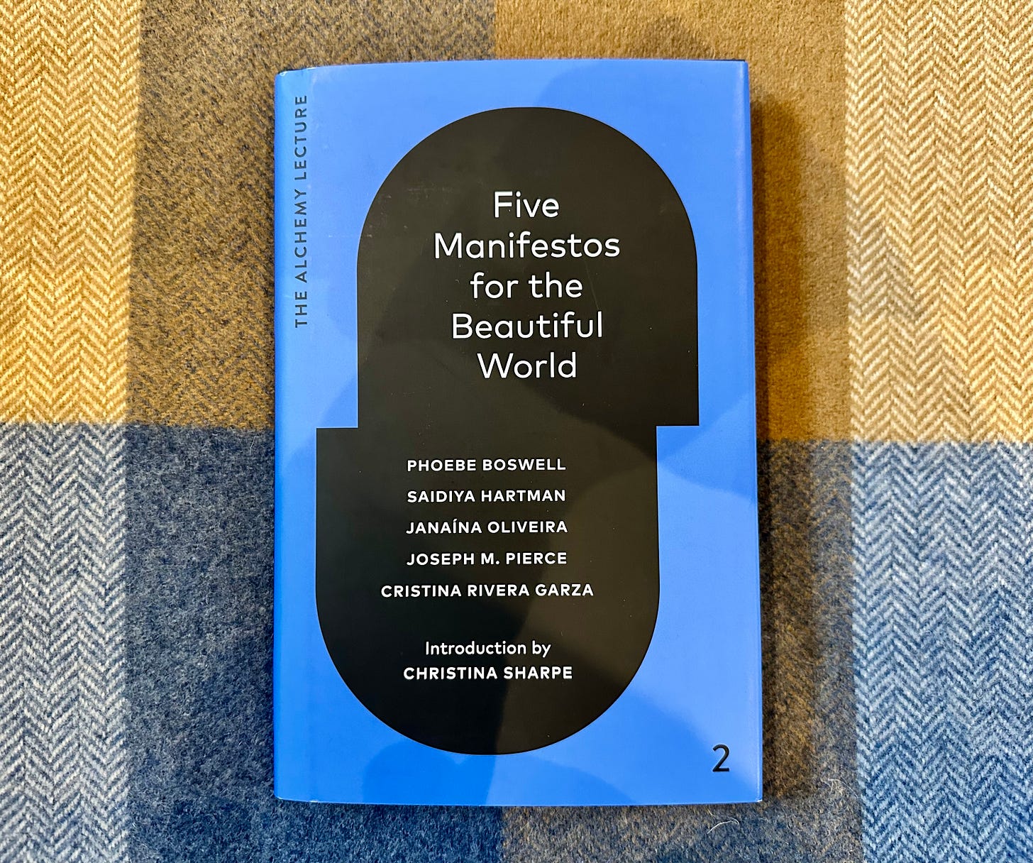 Photo of the book "Five Manifestos for the Beautiful World" drawn together by Christina Sharpe. The book is on a brown and blue herringbone wool blanket.