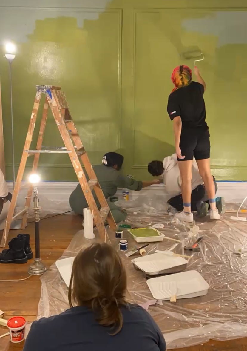Kiana and two of her sisters painting a living room wall green as her sister in law looks on 