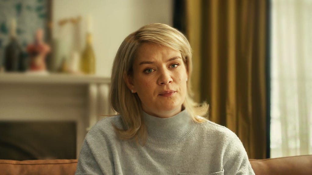 Blonde-haired woman wearing a grey turtleneck jumper sitting on the sofa looking at the camera 