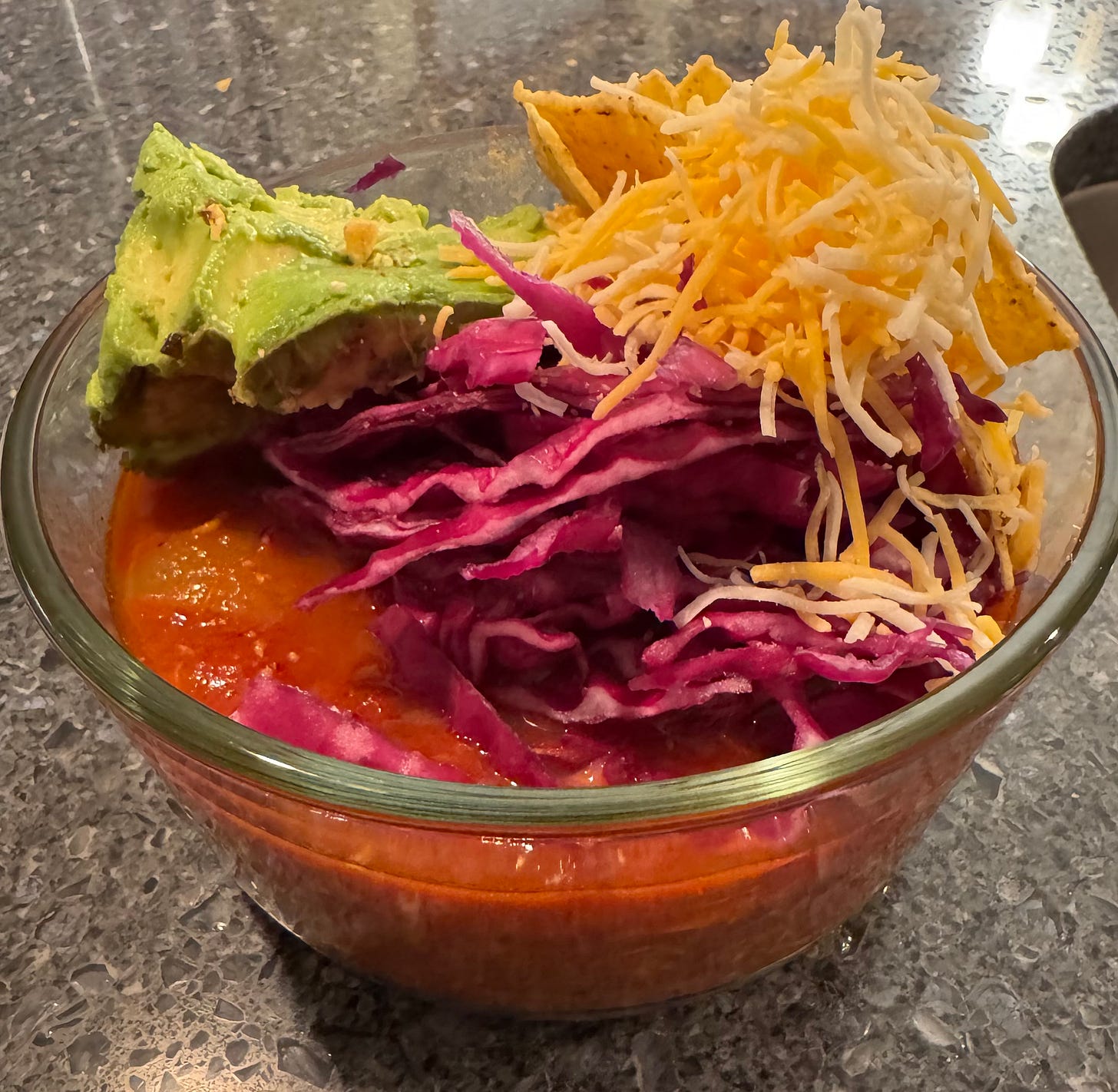 a glass bowl of mediterranean fish soup with 1/2 an avocado, shredded cabbage, tortilla chips, and shredded cheese.