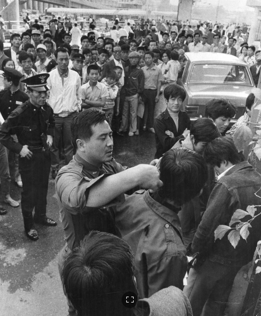 A black-and-white photograph taken in 1971 showing a uniformed and plainclothes South Korean police officer forcibly cutting the hair of a young man with scissors on a public street near Seoul Station. A crowd of onlookers and several police officers stand nearby. This image captures the government's aggressive crackdown on 'improper' hairstyles (long hair on men) and dress codes during the Park Chung-hee authoritarian era.