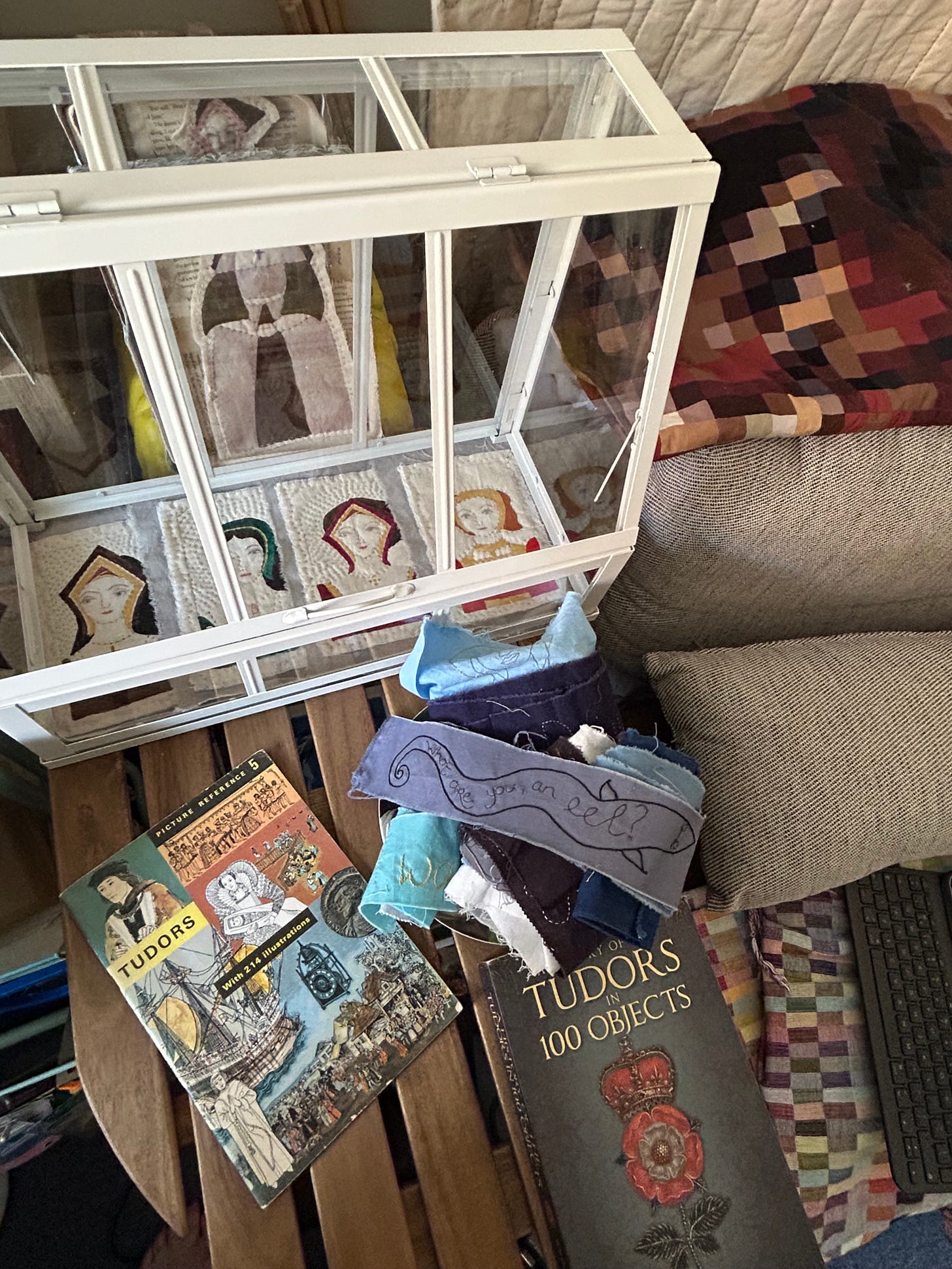 A table top with two books about the Tudors, a pile of rolled stitches with an eel visible on the top. A tabletop greenhouse containing images of four of Henry VIII’s queens.