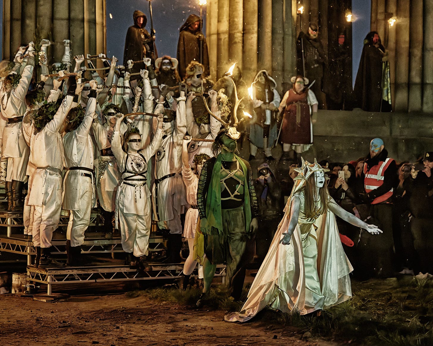 This documentary fieldwork photograph shows a member of May Queen, followed by the Winter Green Man and her attendant protectors. Beltane, Edinburgh 2026.
