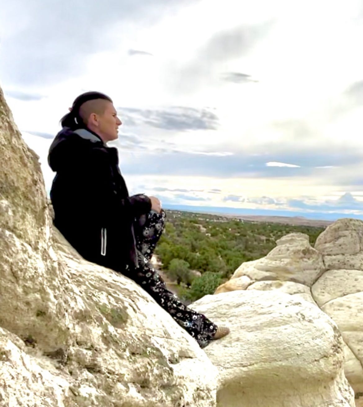 Lyric sitting on the top of the sandstone rocks in the NM dessert. They are wearing black and silver pants, with stars on it, brown boots, and a jacket, looking out over the desert before them. Lyric sitting on the top of the sandstone rocks in the NM dessert. They are wearing black and silver pants, with stars on it, brown boots, and a jacket, looking out over the desert before them.