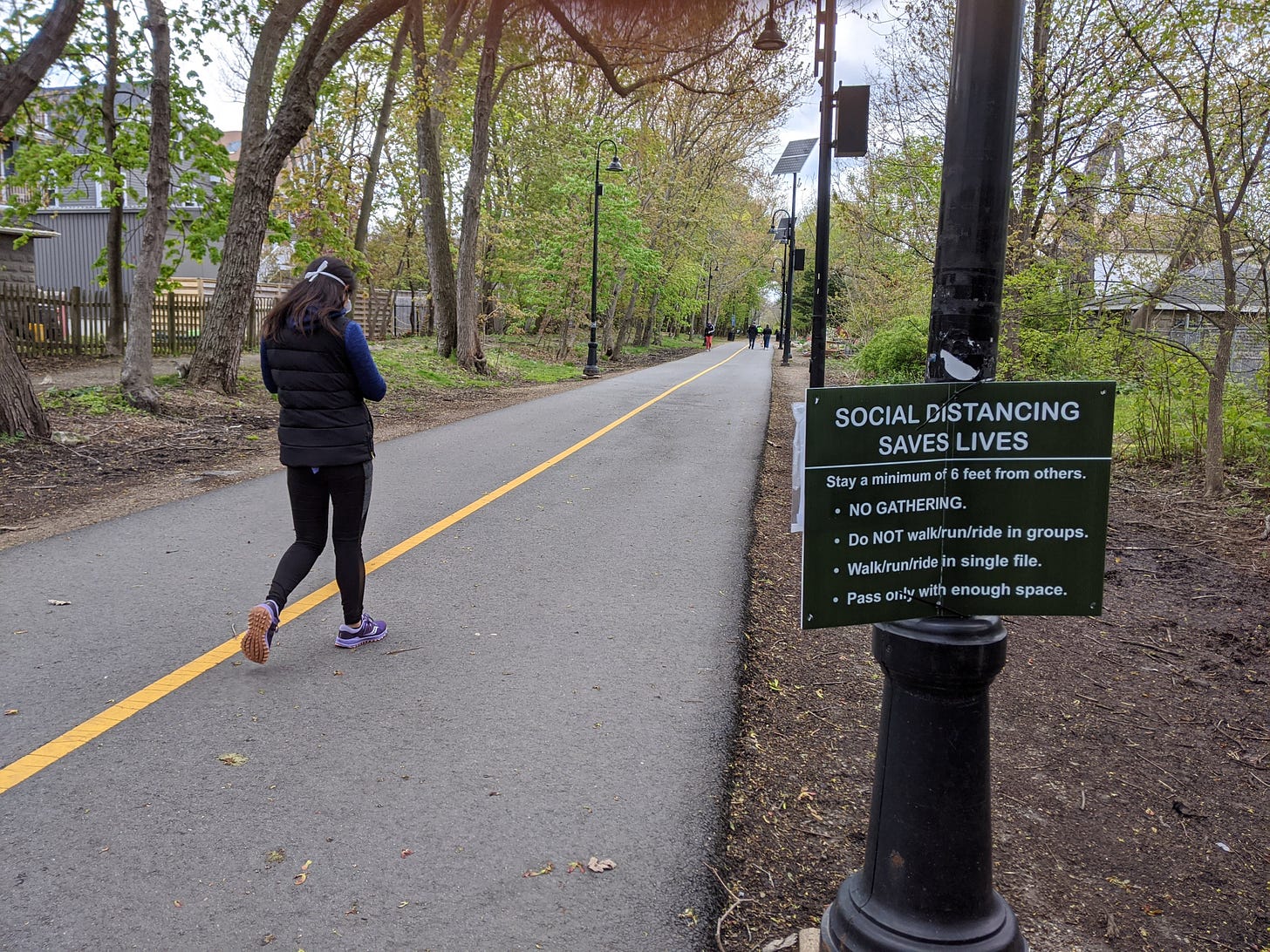 A photograph of an outdoor bike path with a woman walking wearing a mask and a sign reading "SOCIAL DISTANCING SAVES LIVES" among other text in the foreground