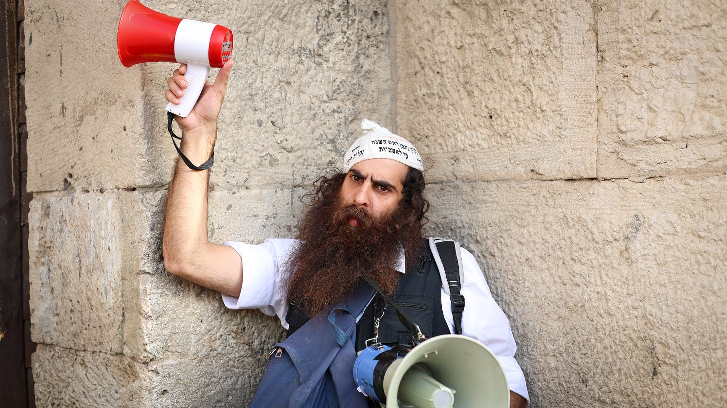 A bearded man with a serious expression leans against a stone wall in Jerusalem, holding a red and white megaphone above his head and wearing another blue megaphone strapped across his chest. He wears a white shirt, black vest, backpack, and a white cloth head covering with Hebrew writing.